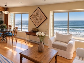 A room with a view of the ocean, a dining area with wooden chairs, a white sofa, and a table with a vase of flowers. - Beach Front Vacation Rental-The Beach is your back yard - Santa Cruz (Watsonville)