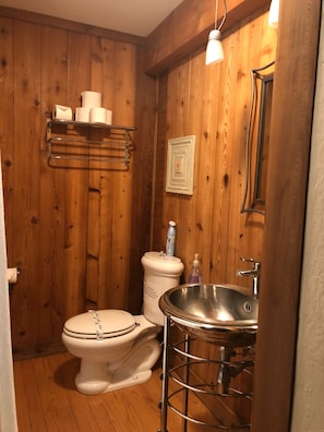 A compact wooden bathroom with a toilet, sink, and a shelf with toiletries.