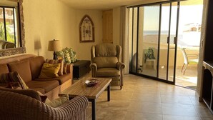 A spacious living room with a large sliding glass door leading to a balcony. There is a brown sofa, a wicker armchair, and a wooden coffee table. - Terrasol 2BR/2BA Beach Condo - Best Price, Best Location & Ocean beachfront (Cabo San Lucas)
