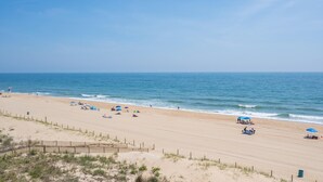 A wide sandy beach with a wooden fence in the foreground, a clear blue sky, and calm ocean waves.