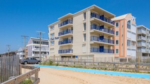 A multi-story building with balconies, a wooden fence, and a sandy beach area.