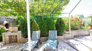Een terras buiten met stenen vloer, een metselwerk open haard en twee loungestoelen onder een pergola.