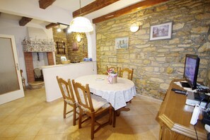 A dining area with a stone wall, wooden chairs, a round table with a white tablecloth, and a television on a wooden stand.