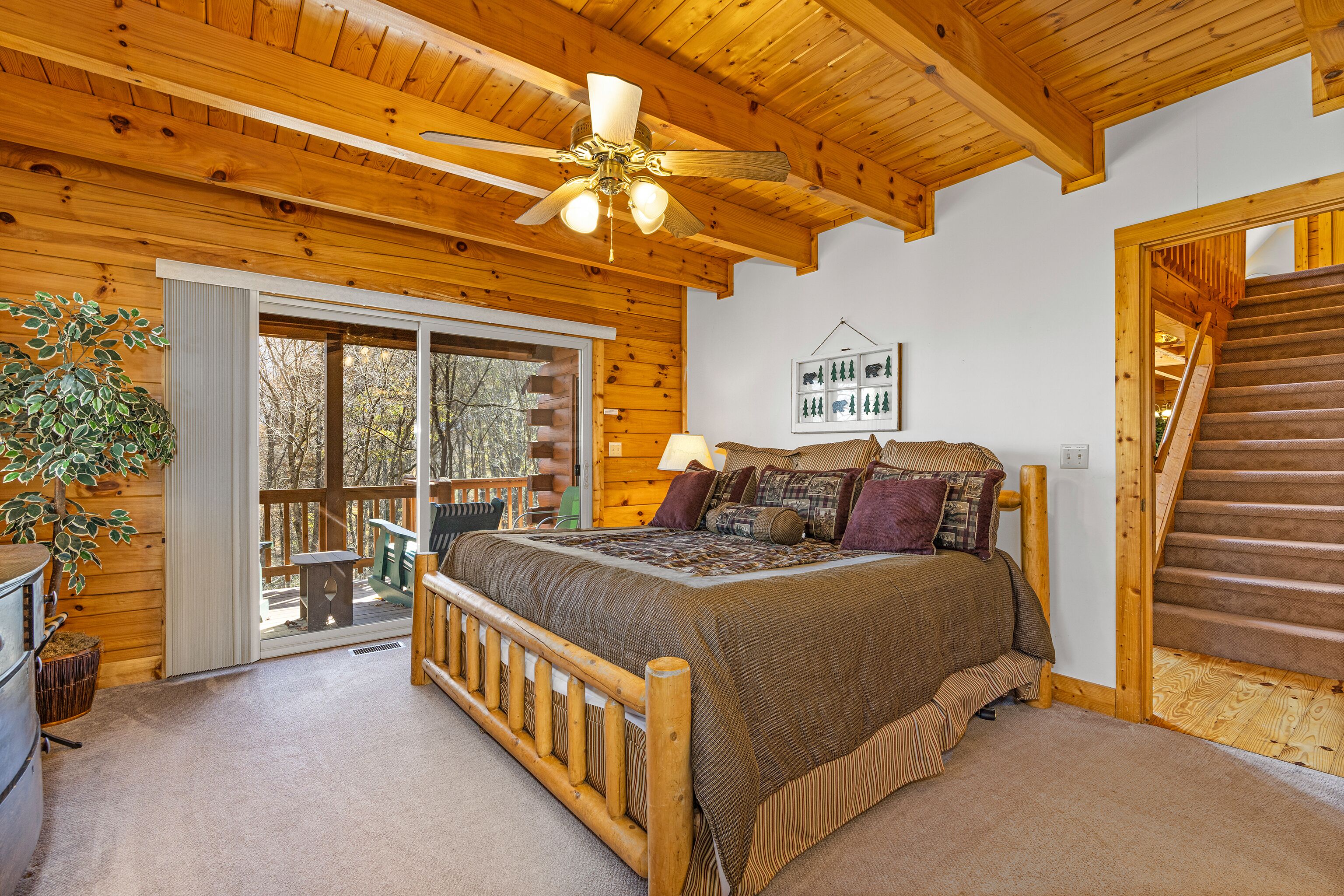 A wooden cabin bedroom with a bed, a ceiling fan, a window, and a staircase.