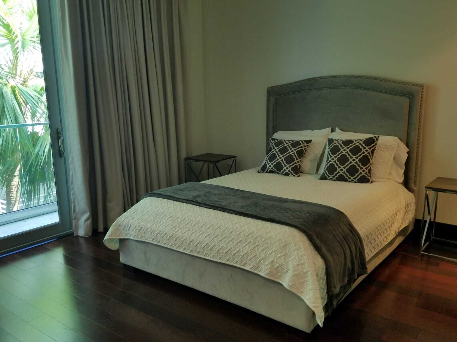 Un letto rifatto con una testiera grigia, un comodino e vista su palme attraverso una finestra con tende.
