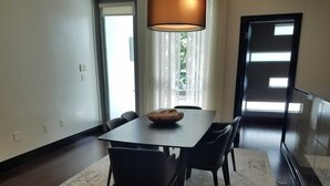 Un comedor con una mesa rectangular, seis sillas, un cuenco de madera, una lámpara de pie grande y vistas al exterior a través de una ventana con cortinas translúcidas.