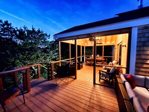 An outdoor deck with a wooden floor, a seating area with chairs and a table, and a glass enclosure leading to a room with a ceiling fan and a window. - Spacious Contemporary, Walk to Private Tru-Haven Beach (Truro)
