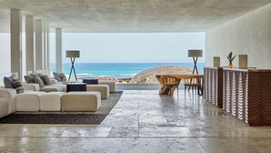 Een moderne woonkamer met een grote bank, een salontafel en een eethoek met stoelen en een tafel. Er is uitzicht op het strand en de oceaan door grote ramen.