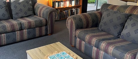 A living room with two patterned sofas, a coffee table, a bookshelf, and a large window with blinds.
