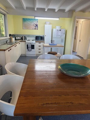Une cuisine de taille réduite, avec des appareils électroménagers blancs, une table à manger en bois et une vue sur le couloir.