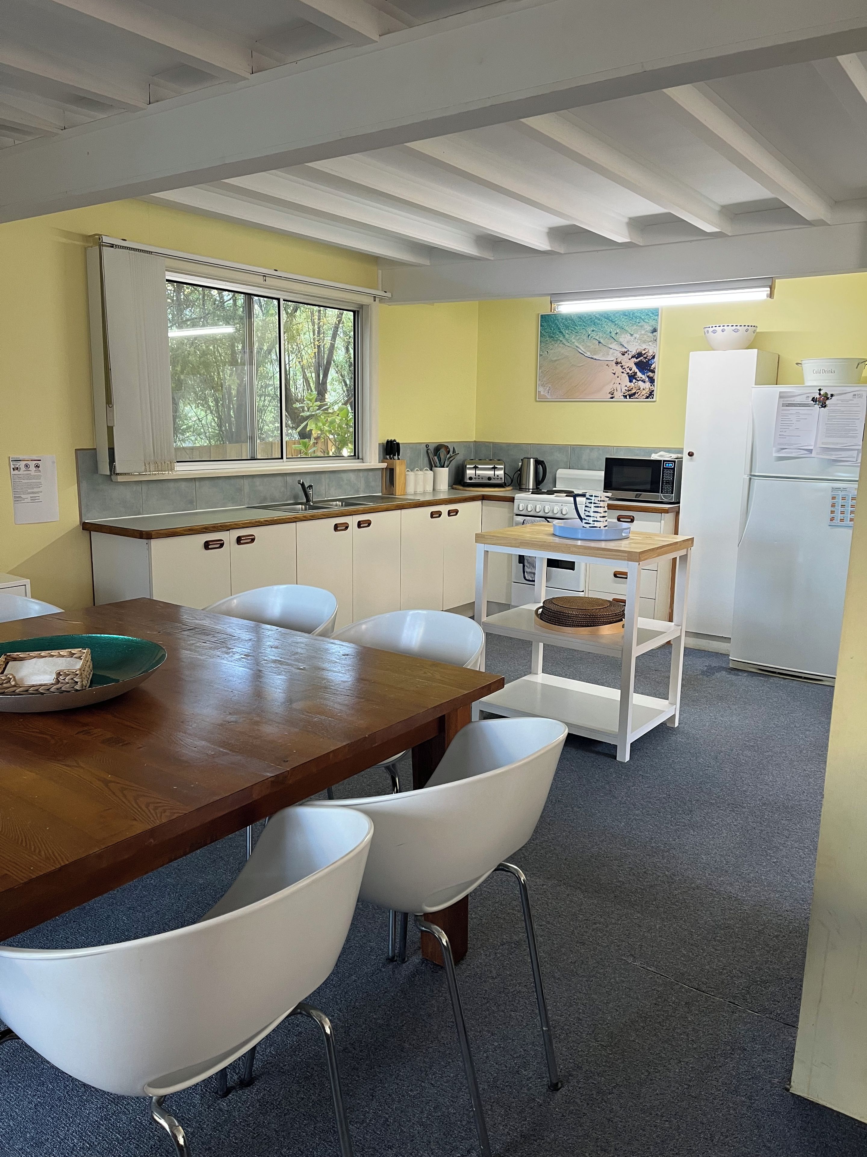 A compact kitchen with a wooden table, white chairs, a microwave, a refrigerator, and a window with a view of trees.