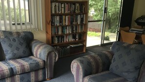 Een woonkamer met twee gestreepte banken, een houten salontafel met een spelletje drie-op-een-rij, een boekenplank vol boeken en uitzicht op groen buiten het raam.