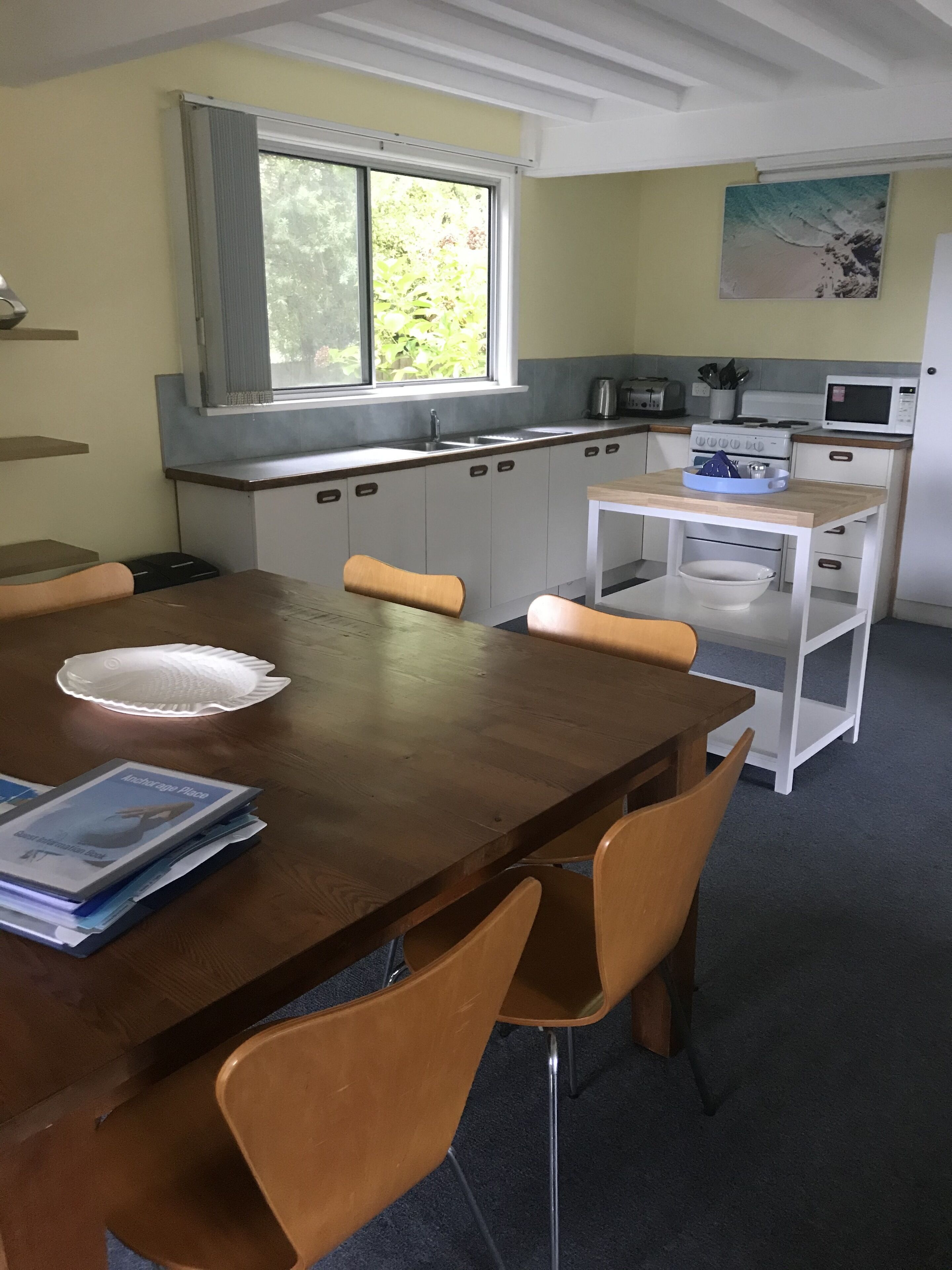 Een eethoek met een houten tafel en stoelen, een keuken op de achtergrond met een raam, en een schilderij aan de muur.