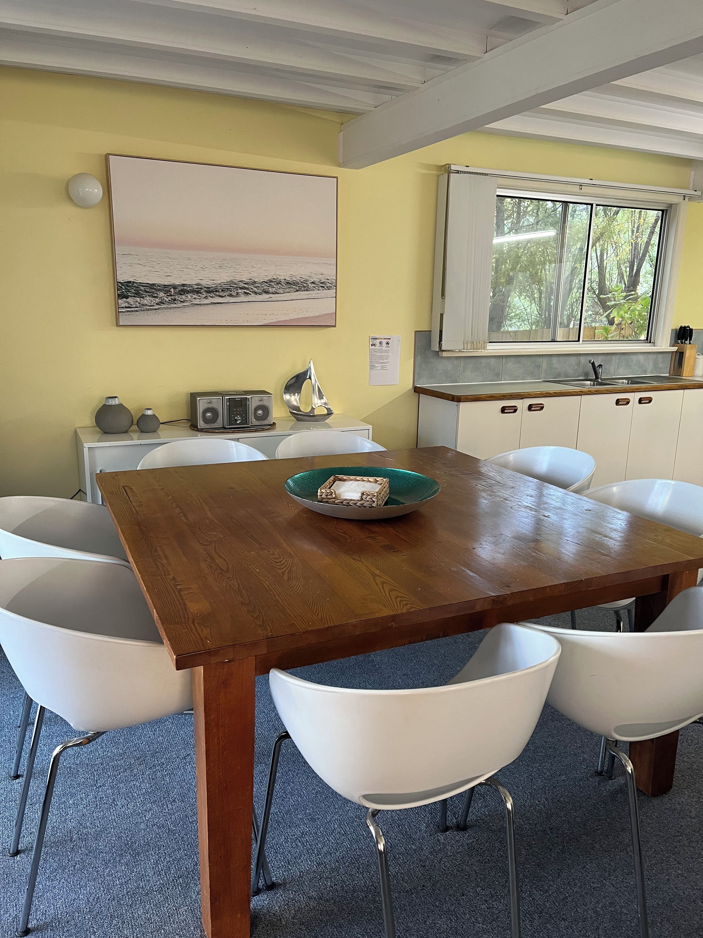 A dining area with a wooden table, white chairs, a bowl of fruit, a radio, and a painting of a beach scene.
