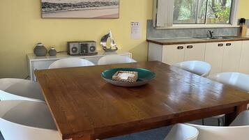 A dining area with a wooden table, white chairs, a bowl of fruit, a radio, and a painting of a beach scene.