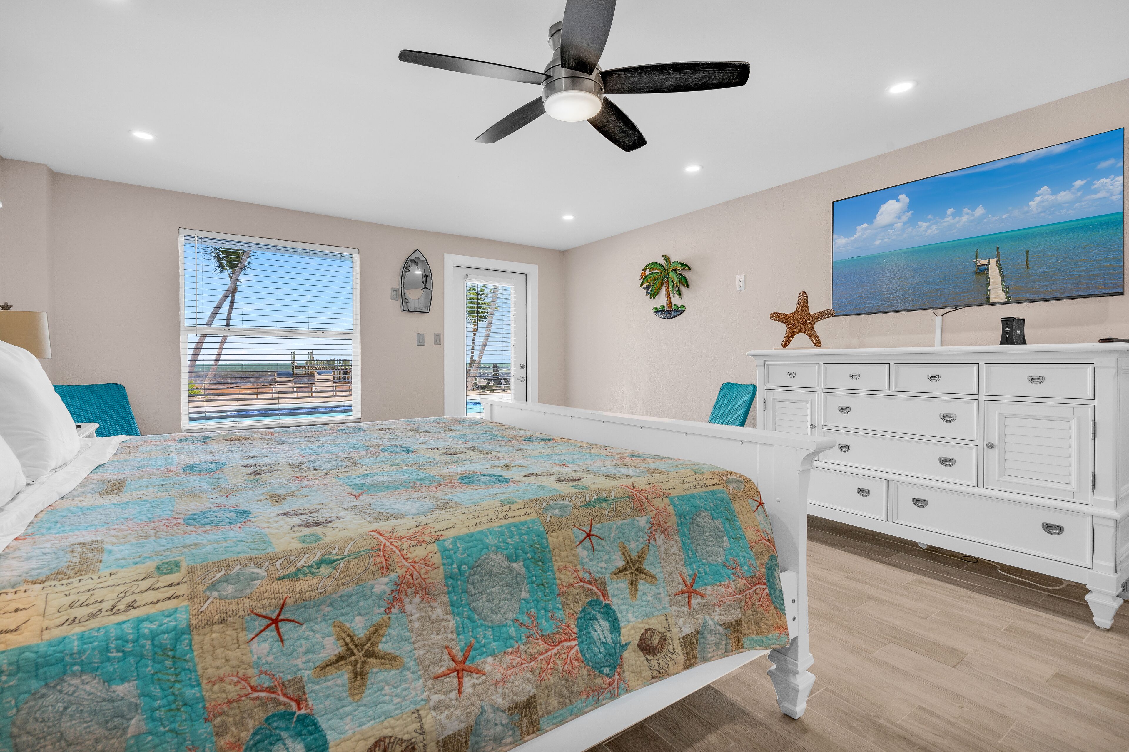 A bedroom with a bed, a ceiling fan, a TV, a dresser, and a view of the ocean through a window.