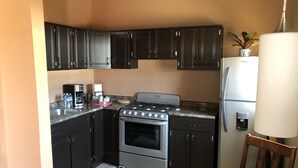 A compact kitchen with dark cabinets, a stove, a refrigerator, and a coffee maker. - Labios Tintos Bungalow, Valle de Guadalupe, BC, México (Las Lomas)