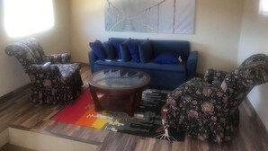 A living room with a blue sofa, two floral-patterned armchairs, a glass coffee table, and a red rug. - Labios Tintos Bungalow, Valle de Guadalupe, BC, México (Las Lomas)