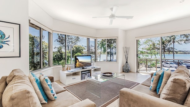 A modern living room with a large TV, comfortable seating, and a view of the sea. - Aarons Villa - Waterfront Perfectly located on Gympie Terrace (NOOSAVILLE)