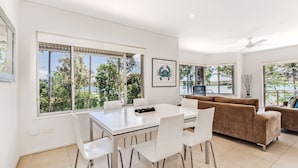 A modern living room with a dining area, large windows, and a view of greenery. - Aarons Villa - Waterfront Perfectly located on Gympie Terrace (NOOSAVILLE)