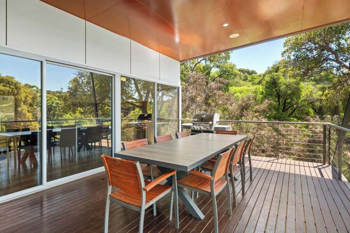 A modern outdoor dining area with a large table and chairs, surrounded by glass walls and a deck overlooking a lush landscape.