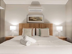 Una habitación de hotel con una cama grande, dos mesitas de noche con lámparas, un aire acondicionado de pared y un cuadro enmarcado sobre la cama.