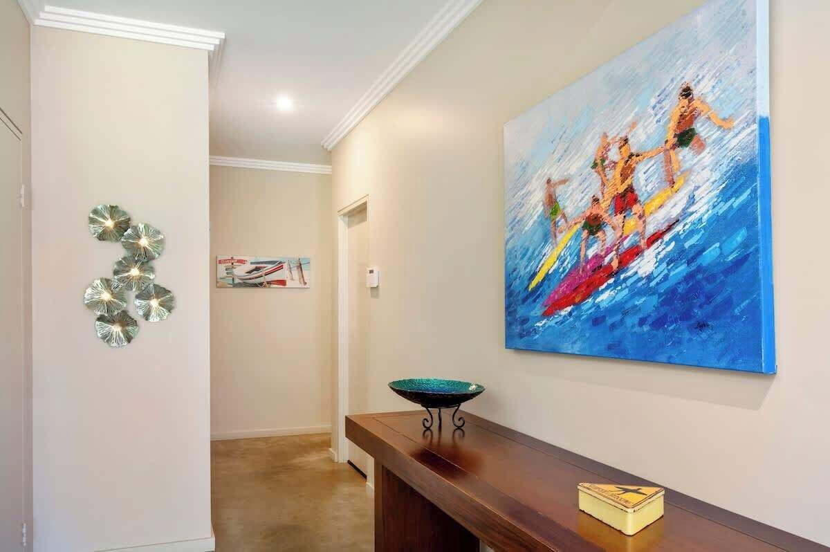A hallway with a painting of surfers, a decorative wall piece, and a bench with a bowl.