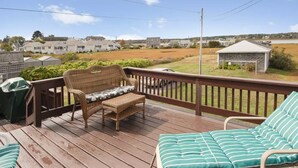 Une terrasse en bois, agrémentée de meubles en osier et d’une chaise en métal, qui offre une vue sur un quartier résidentiel avec ses maisons et un champ verdoyant.