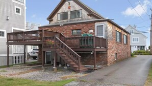 Une maison à deux étages, construite en briques, avec une terrasse en bois et un escalier menant à la porte d’entrée.