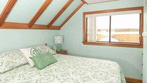 Une chambre avec un plafond en pente, un lit avec une literie à motifs, une table de chevet avec une lampe et une fenêtre donnant sur la plage.