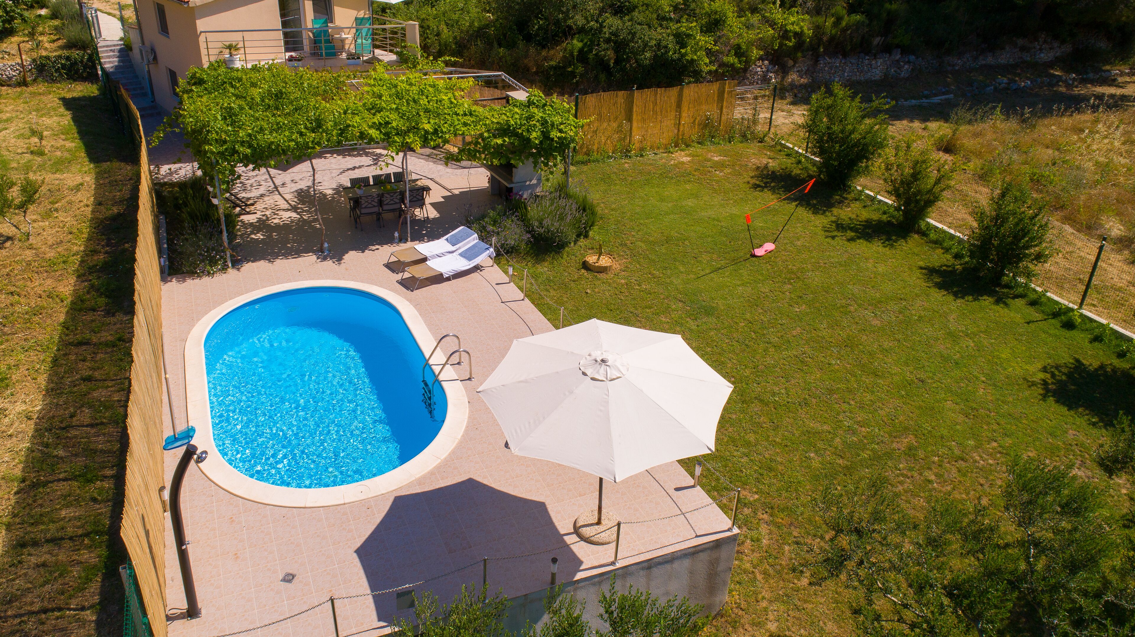 Eine Vogelperspektive zeigt einen Garten mit Schwimmbad, einer Terrasse mit Stühlen und Tisch sowie einen eingezäunten Bereich mit einer Schaukel.