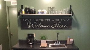 A compact kitchen with a black refrigerator, a sink, and a coffee maker. Above the sink, there is a shelf with jars and a sign that reads "LOVE LAUGHTER & FRIENDS Welcome Here."
