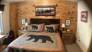 A cozy room with a wooden log wall, a bed with a bear-themed bedspread, two bedside tables with lamps, and a framed picture above the bed.