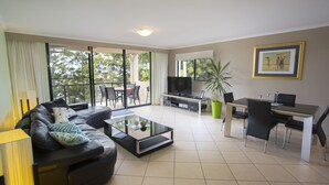 A modern living room with a sofa, coffee table, TV, and dining area. - Oceanview 6 at Nambucca with rooftop tce & spa (Nambucca Heads)