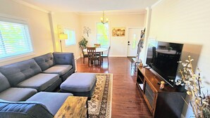 A living room with a grey sofa, a coffee table, a TV, and a dining area with a table and chairs. - Charming duplex with private yard. Close to the beach, dining & shopping! (Greenwich)