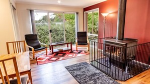 Un salon chaleureux avec une cheminée, deux fauteuils, une table et une vue sur la verdure grâce à de grandes fenêtres.