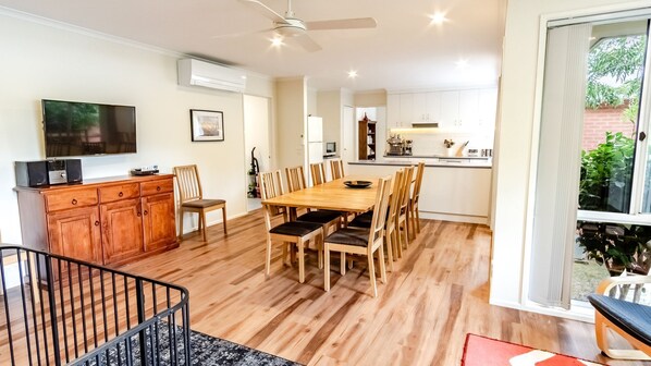 A spacious living area with a wooden dining table and chairs, a flat-screen TV, and a kitchen in the background. - HARGREAVES HOUSE: Bright (Bright)