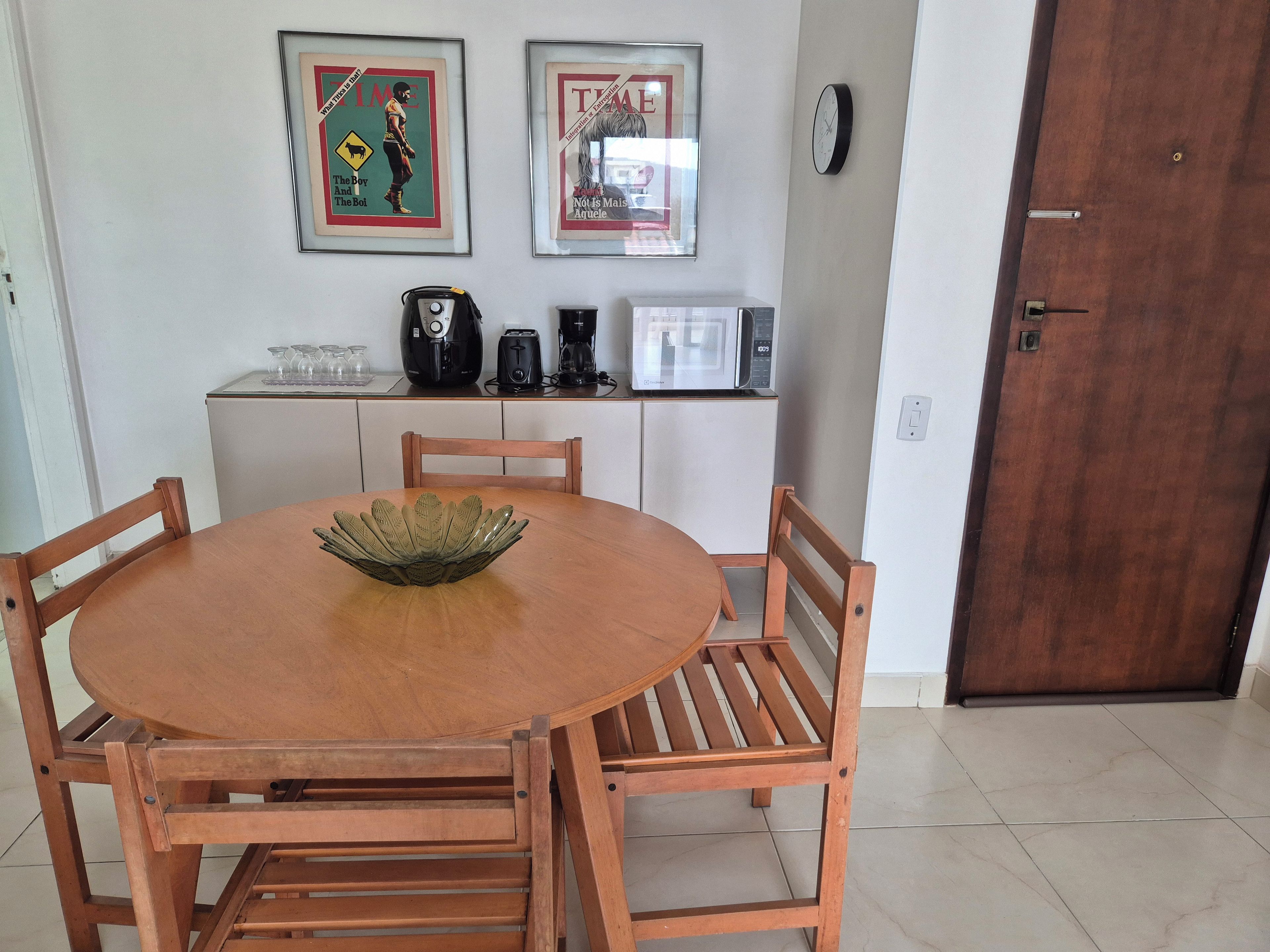 A dining area with a round wooden table, four chairs, a decorative bowl, a microwave, a coffee maker, and two framed Time magazine covers on the wall.
