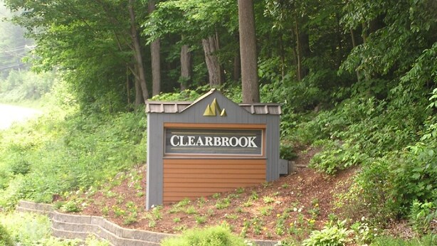 A sign for 'CLEARBROOK' with a wooden garage door, surrounded by greenery and a stone retaining wall.