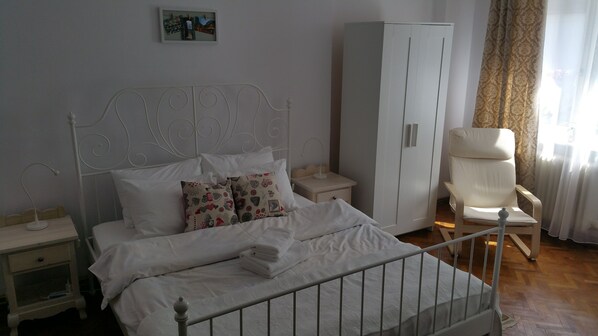 A neatly made bed with white linens and decorative pillows, a bedside table with a lamp, a wardrobe, and a chair in a well-lit room. - Amazing house with beautiful garden, 5 bedrooms, terrace, in the City Centre (Sibiu)