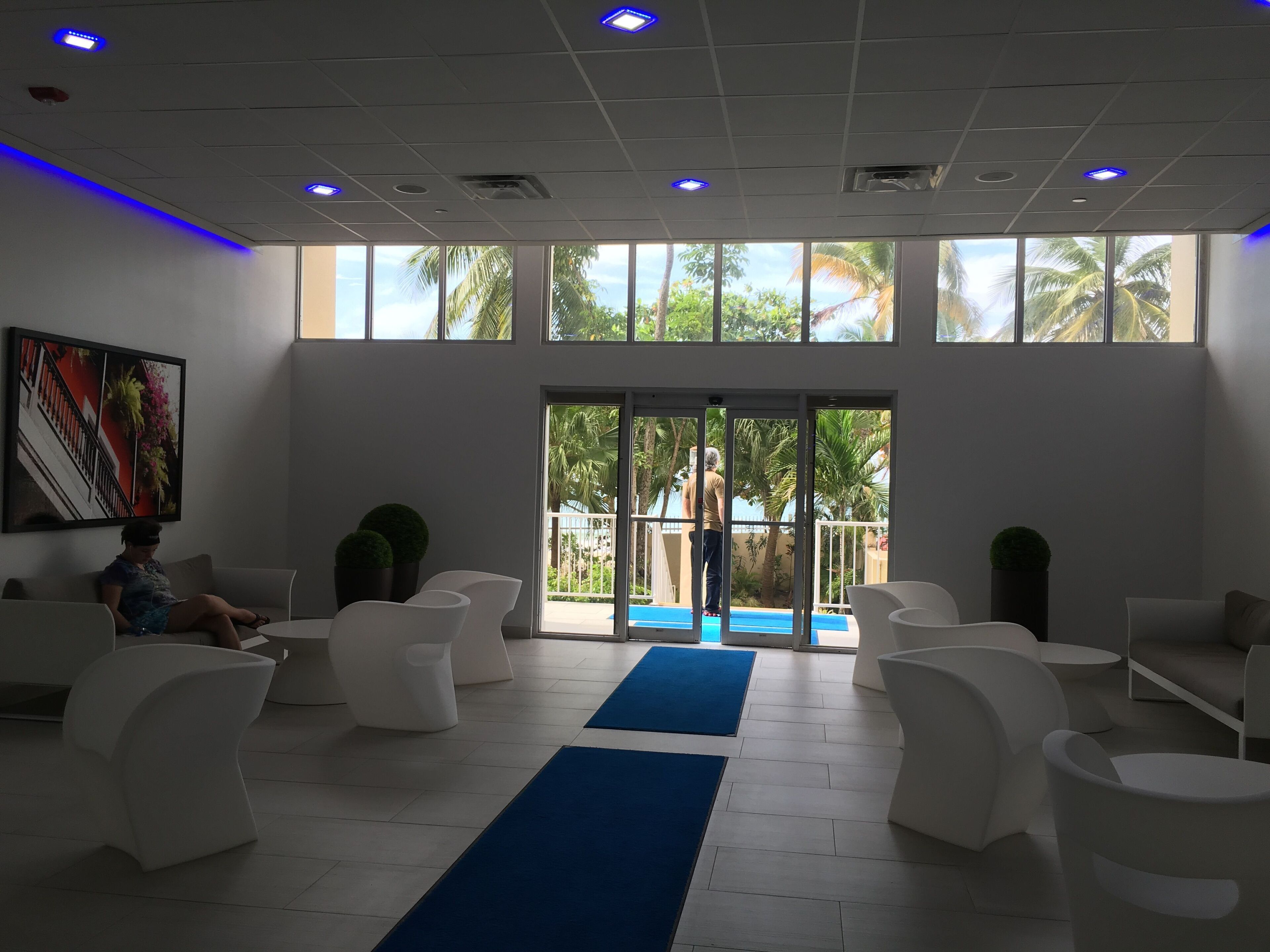 A spacious lobby with a blue carpet leading to a glass door, white chairs, and a large window with a view of palm trees.