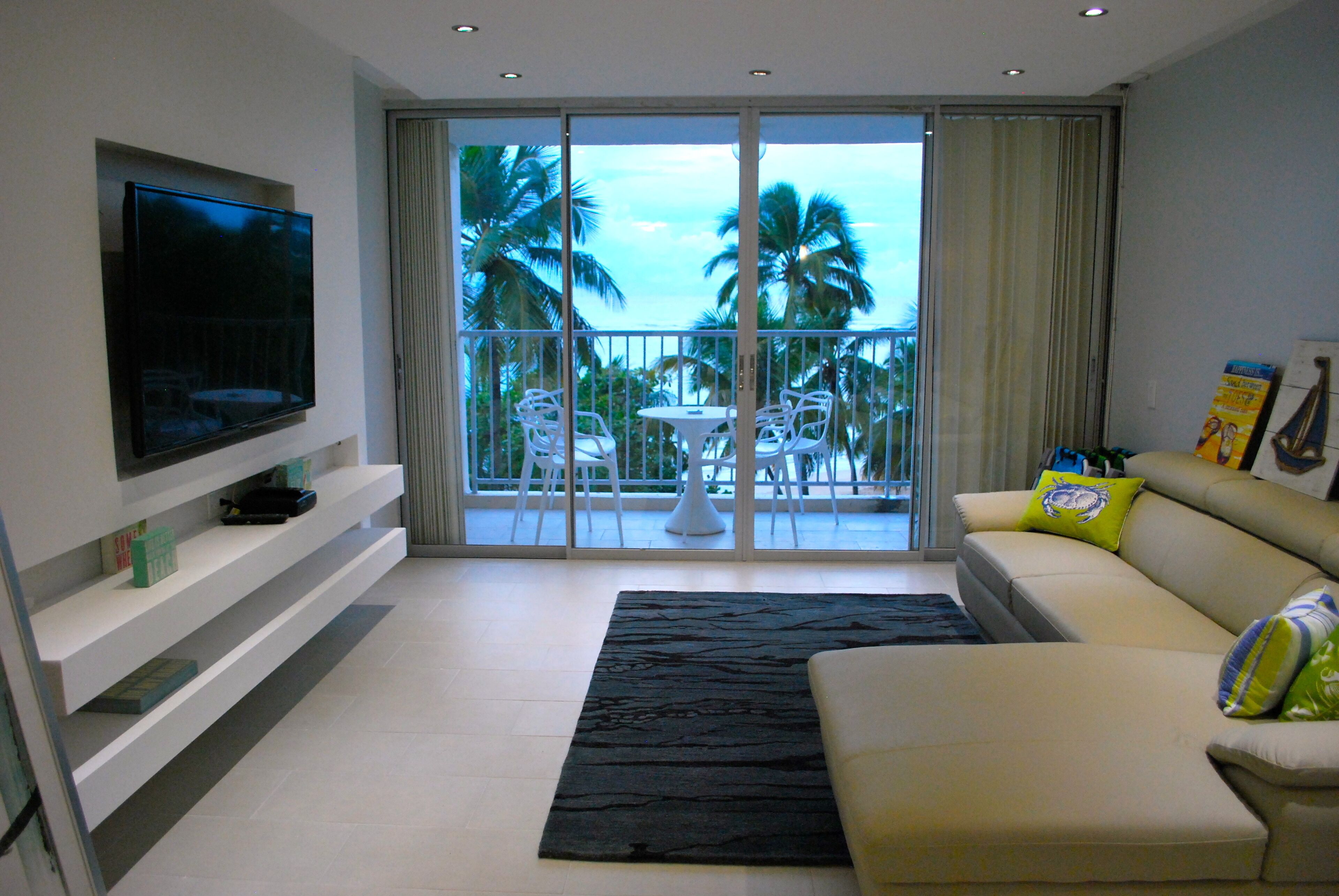 A modern living room with a flat-screen TV, a sofa, a coffee table, and a balcony with a view of palm trees.