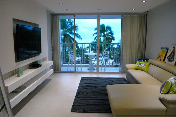 A modern living room with a flat-screen TV, a sofa, a coffee table, and a balcony with a view of palm trees.