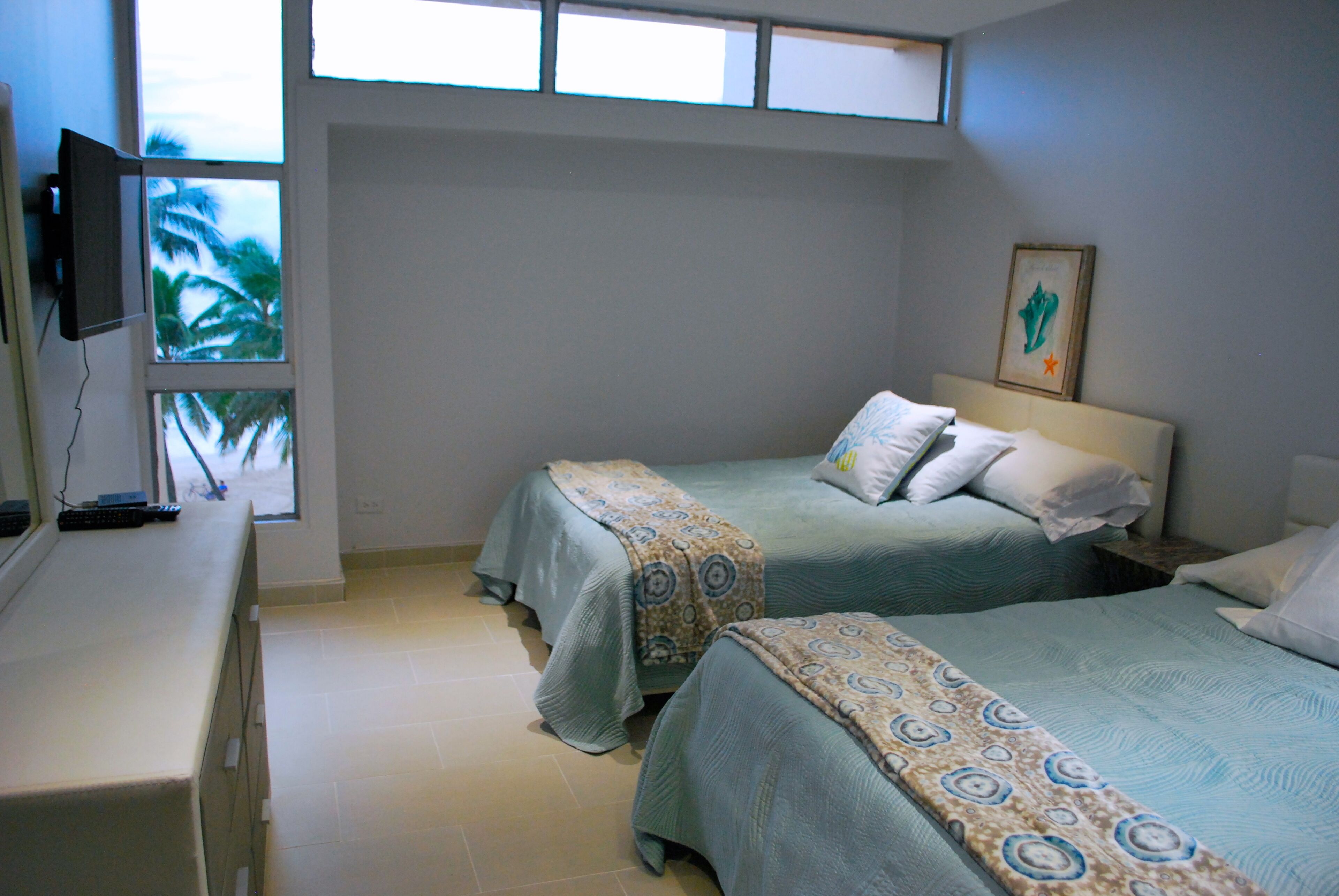 A hotel room with two beds, a TV, and a window with a view of palm trees.