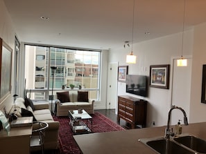 Uma sala de estar moderna com sofá, mesa de centro e TV. Há uma janela grande com vista para edifícios.