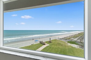 A view of a beach from a window, including the ocean, a grassy area, and a pathway.
