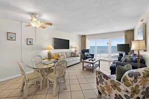 Quarto de hotel com varanda, área de jantar com cadeiras e mesa, sofá, televisão e ventoinha de teto.