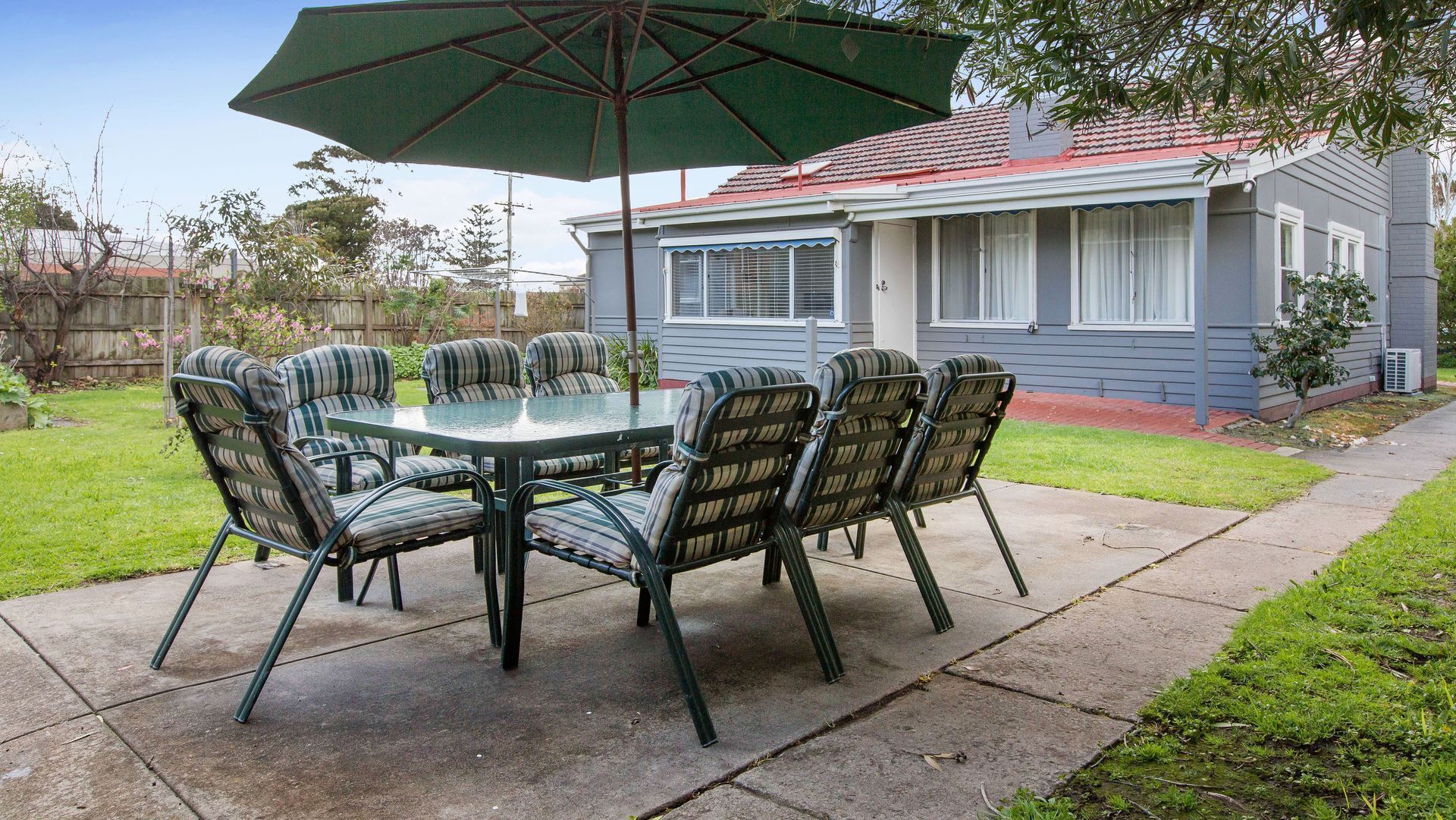 Een terras met een ronde tafel en stoelen, een parasol en een huis op de achtergrond.
