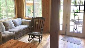 Interior d'una cabana de fusta amb un sofà, una cadira bressol i grans finestres.