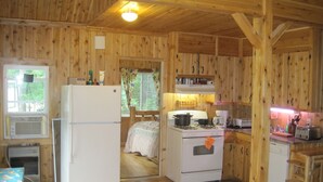 Interior d'una cabana de fusta amb zona de cuina, nevera, fogó i finestra amb cortines.